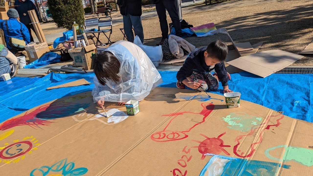 屋外に広げた大きな段ボールに、親子で絵の具を使って絵を描く様子