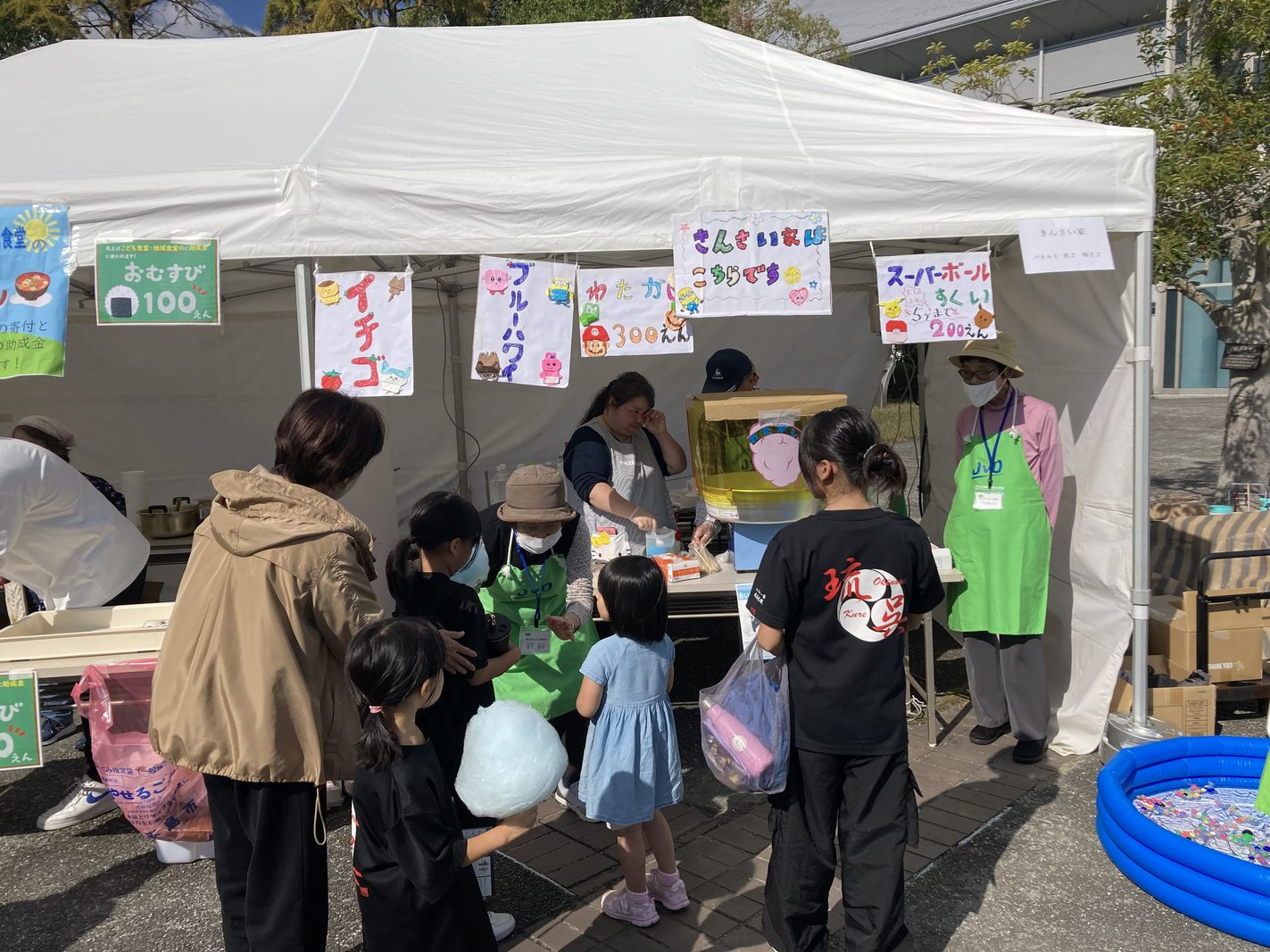 屋外テントの模擬店。手書きのメニューが下がり、子どもたちが綿あめなどを買い求める様子