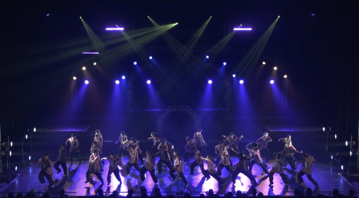 ホールのステージで大勢の出演者が照明を浴びて踊る群舞の場面